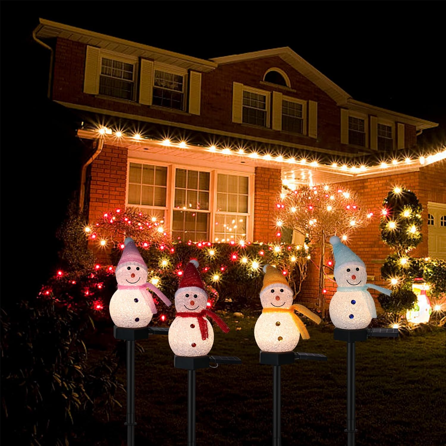 Four solar snowman pathway holiday lights with pink, red, yellow, and blue hats glowing in a row in front of a lit brick house at night