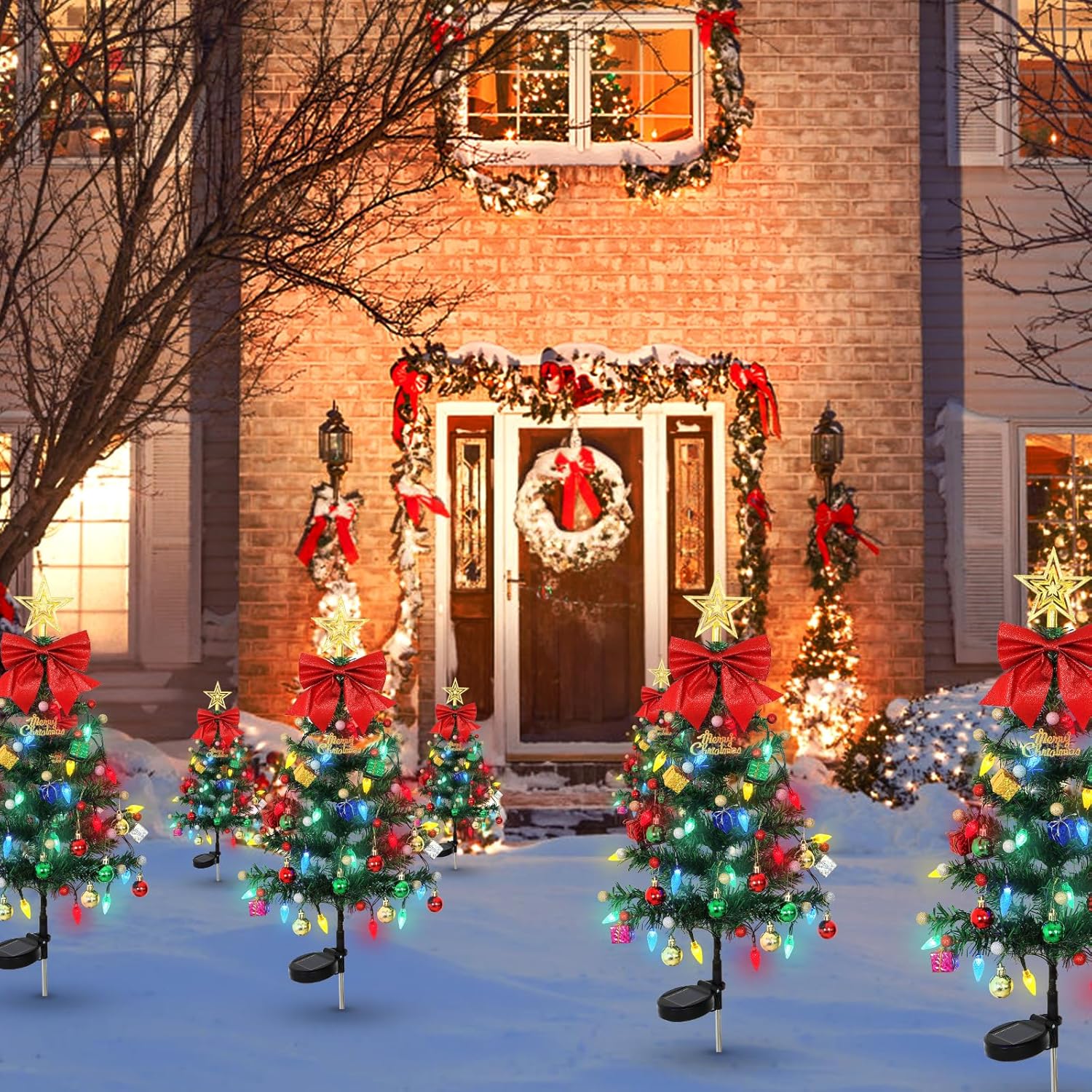 Solar Christmas tree yard lights in a row along a brick home driveway at night, each tree decorated with colorful LED ornaments, red bow, and star topper on ground stakes