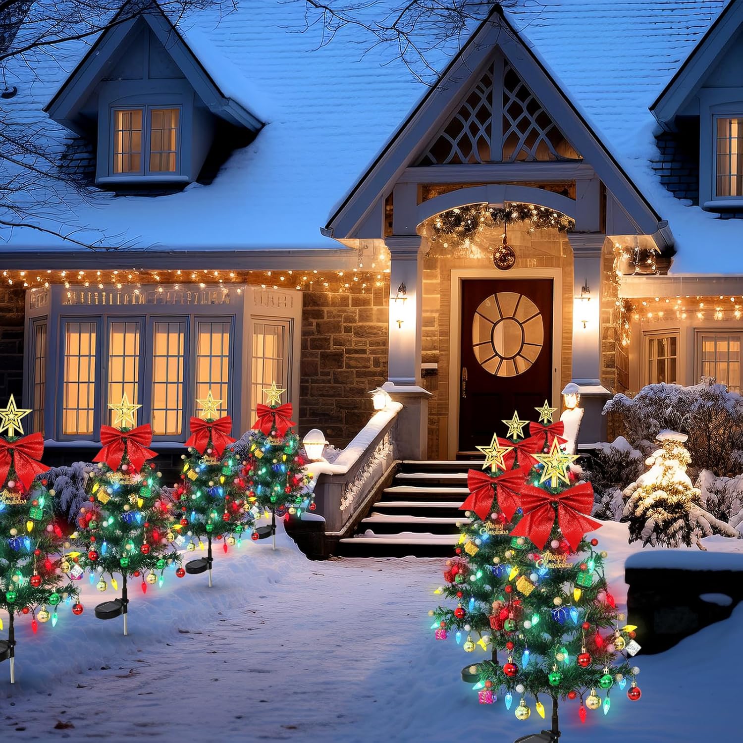 Four decorated solar Christmas tree yard stakes with multicolor ornament lights and red bows lining a stone front walkway beside a lit traditional home at night in light snowfall