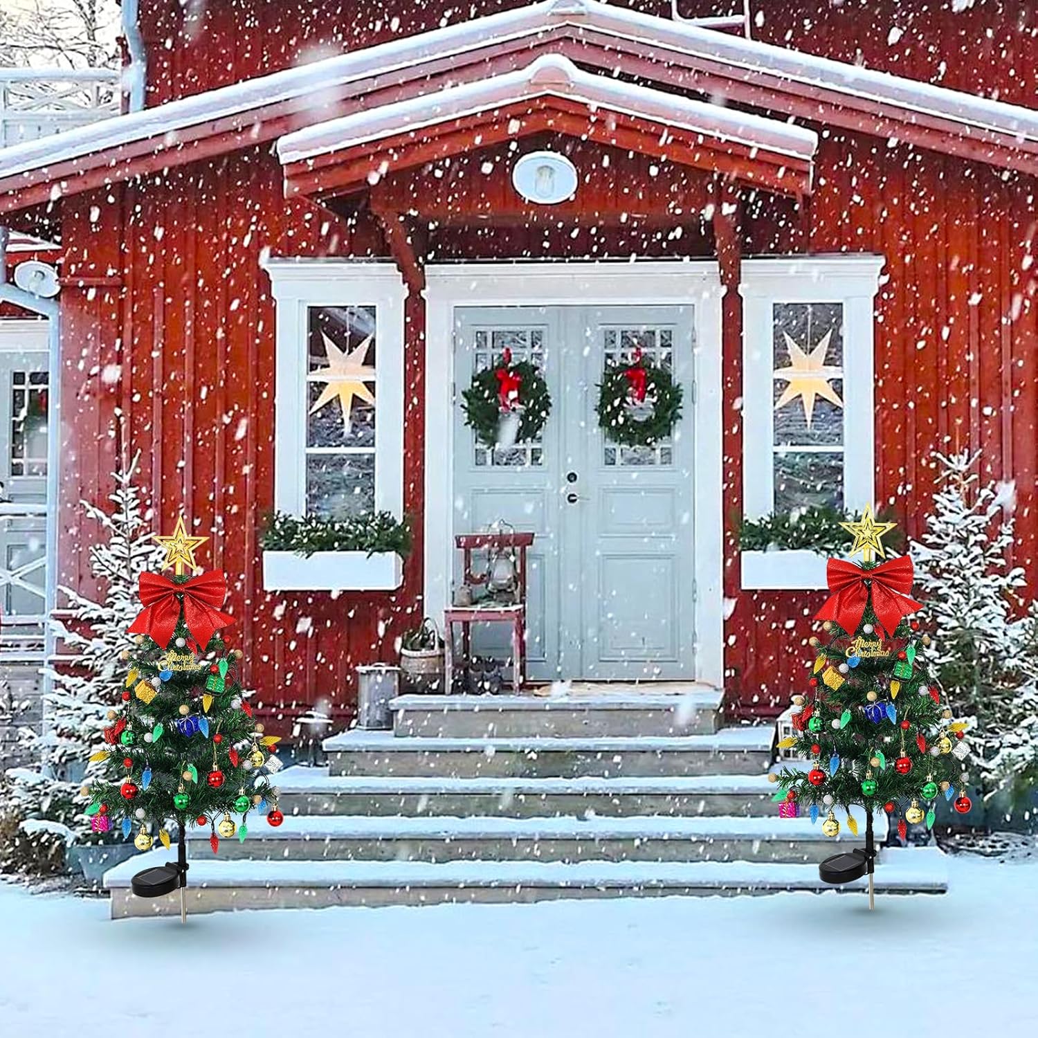 Two solar Christmas tree yard decorations flanking the front steps of a red wood-sided home in snow, each tree with red bow and multicolor LED ornaments on ground stakes