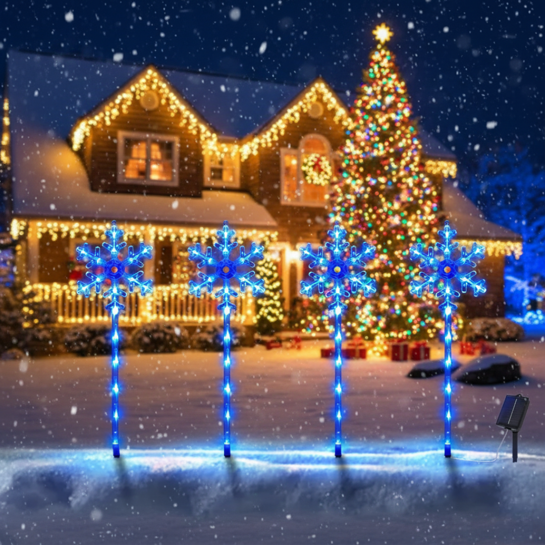 Set of 4 solar snowflake pathway lights with blue and amber LED accents staked in snow in front of a warm-lit holiday home and Christmas tree