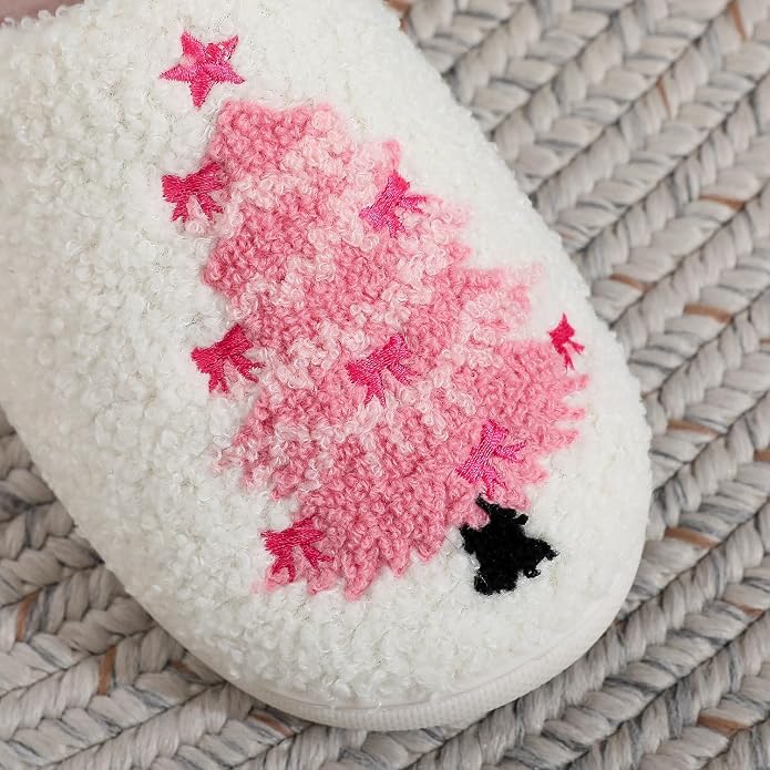 Close-up of a white fluffy slipper with a textured sherpa-like surface, featuring an embroidered pink Christmas tree design with a hot pink star topper and small black trunk detail, resting on a woven surface.