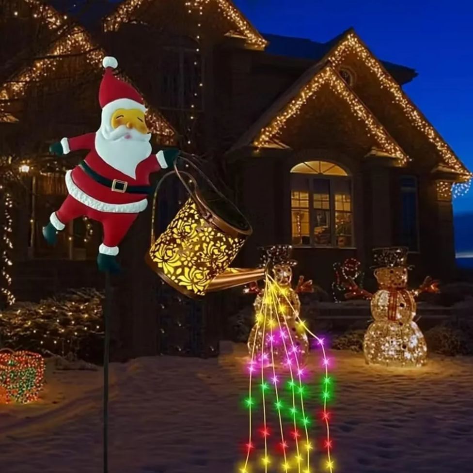 Solar Santa kettle garden stake light in a snowy yard with a lit log cabin backdrop, copper watering can glowing and spilling warm LED strings
