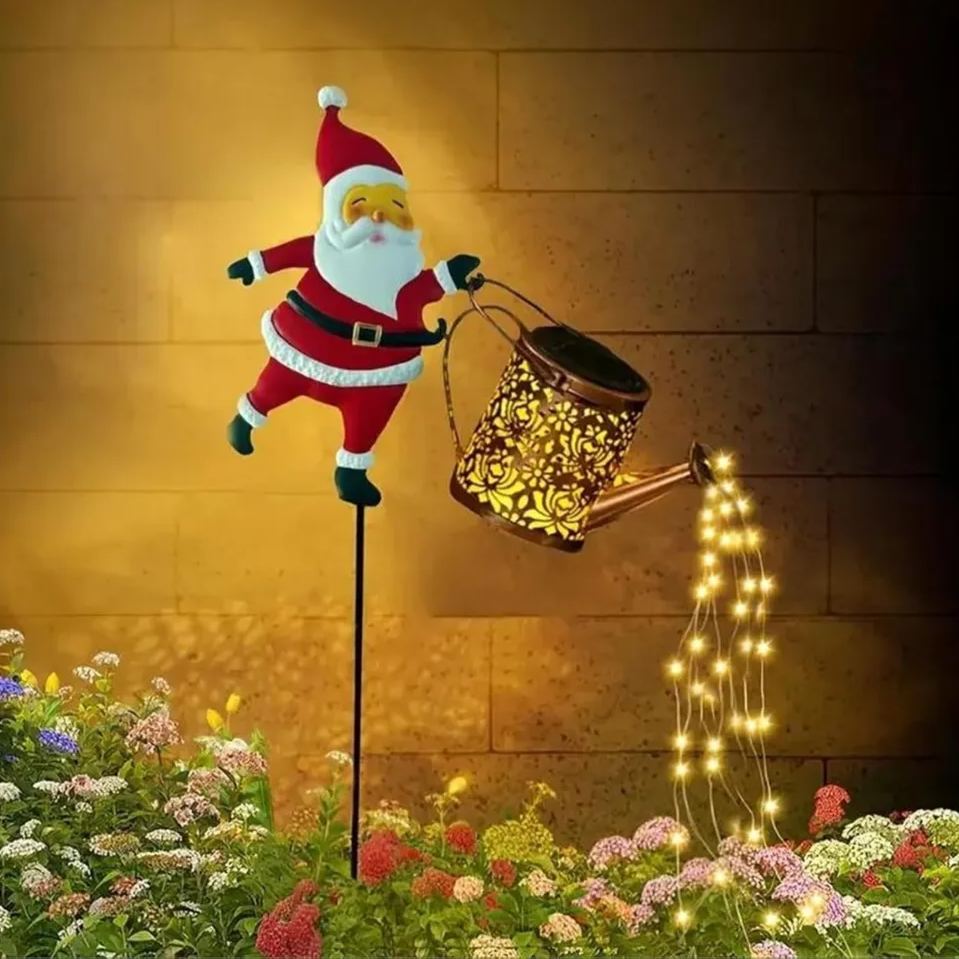 Solar Santa kettle garden stake light against a stone wall with warm-white LED strings cascading from the copper watering can into a flower bed