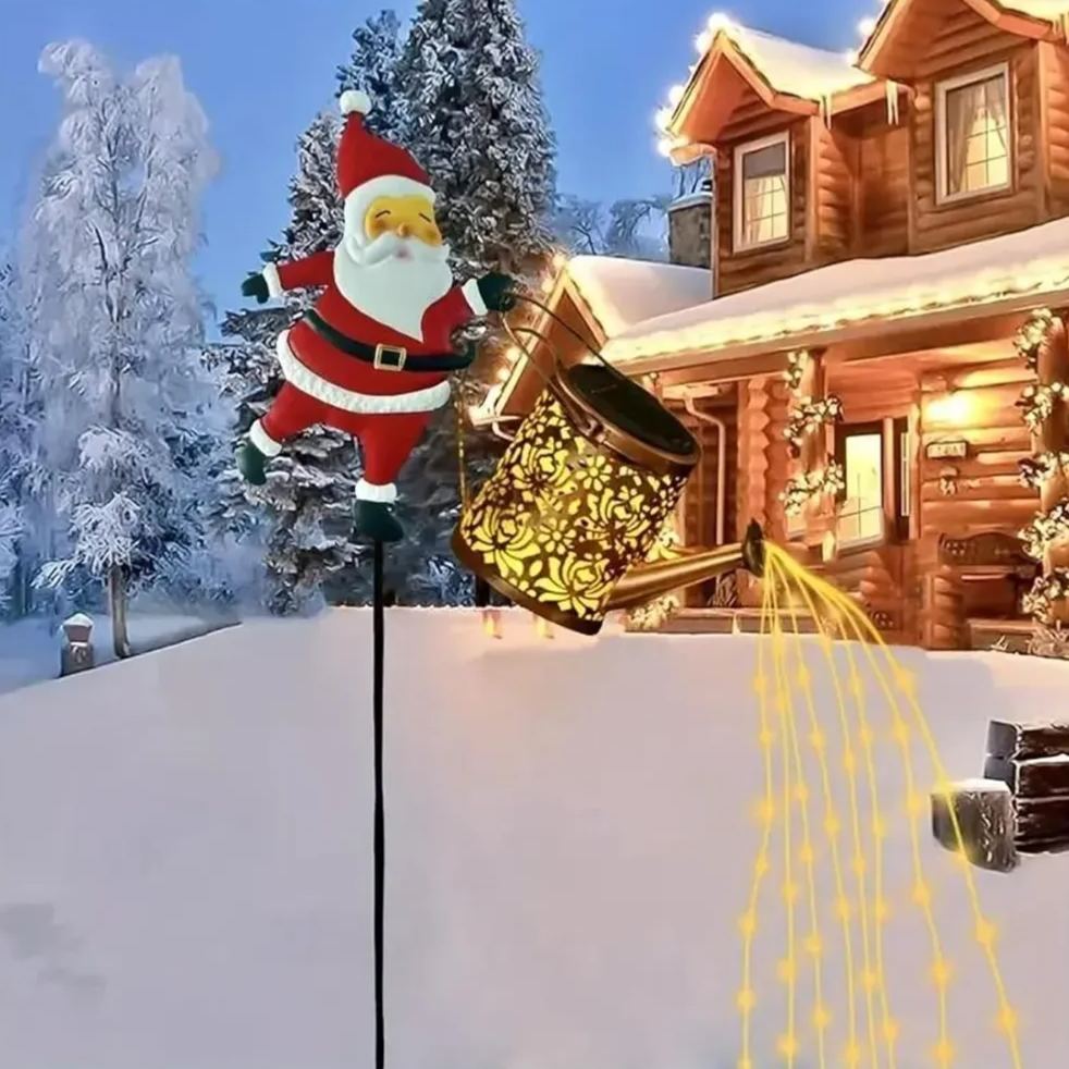 Solar Santa kettle garden light stake in a snowy winter yard with a log cabin background, warm-yellow LED strings flowing from the tilted watering can