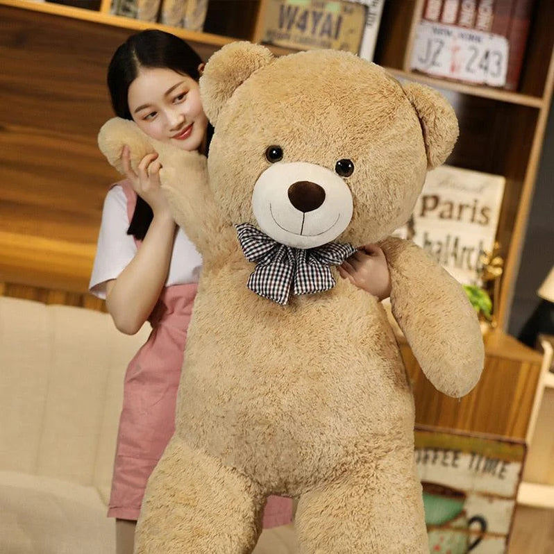 A woman playfully posing with a beige giant teddy bear wearing a checkered ribbon.