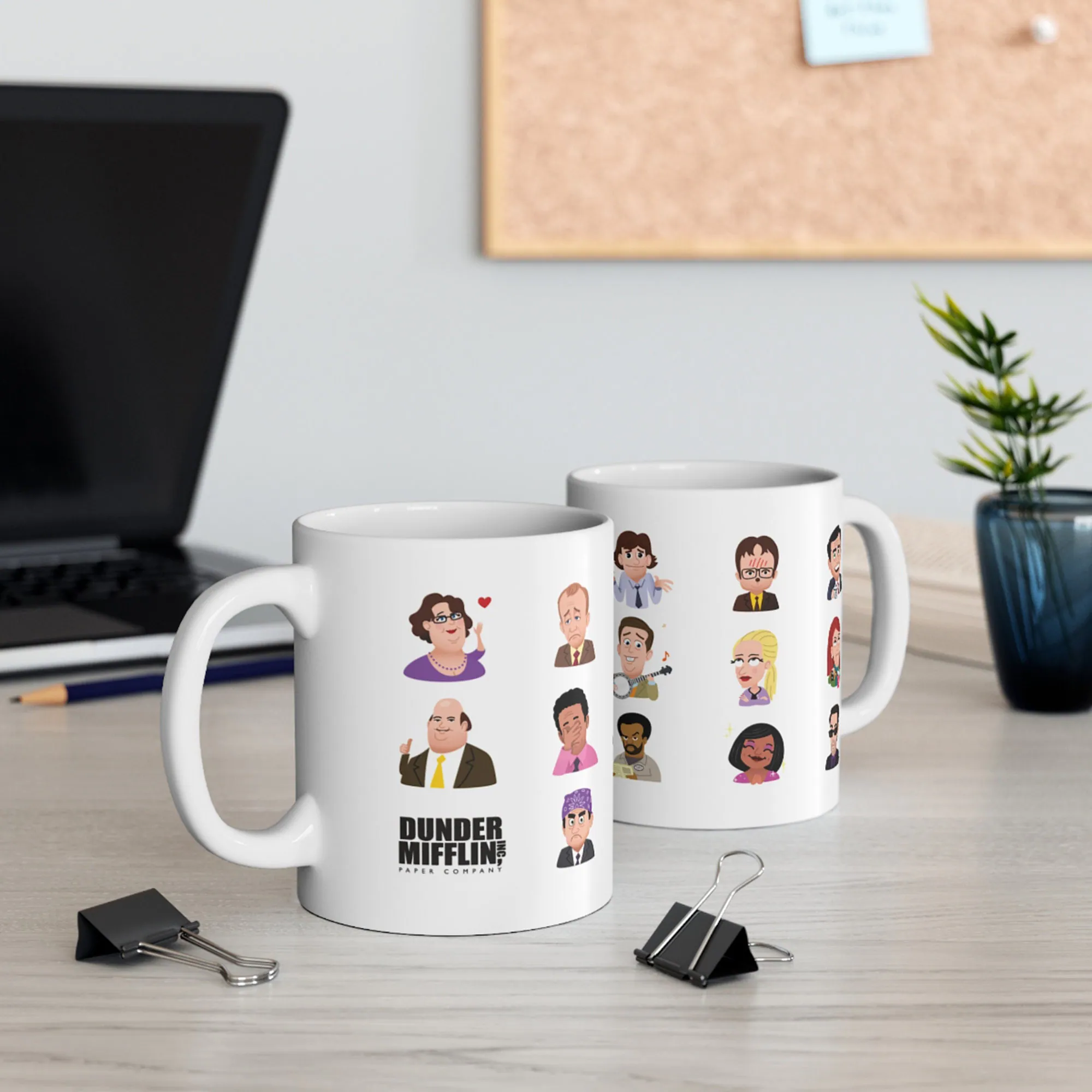 Two white ceramic mugs with various character portraits and Dunder Mifflin branding on a desk