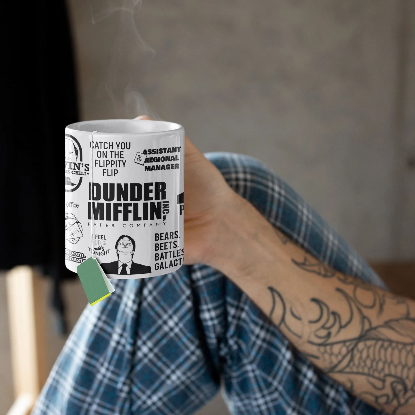 Person holding a white Dunder Mifflin themed coffee mug in bed with a tea bag inside