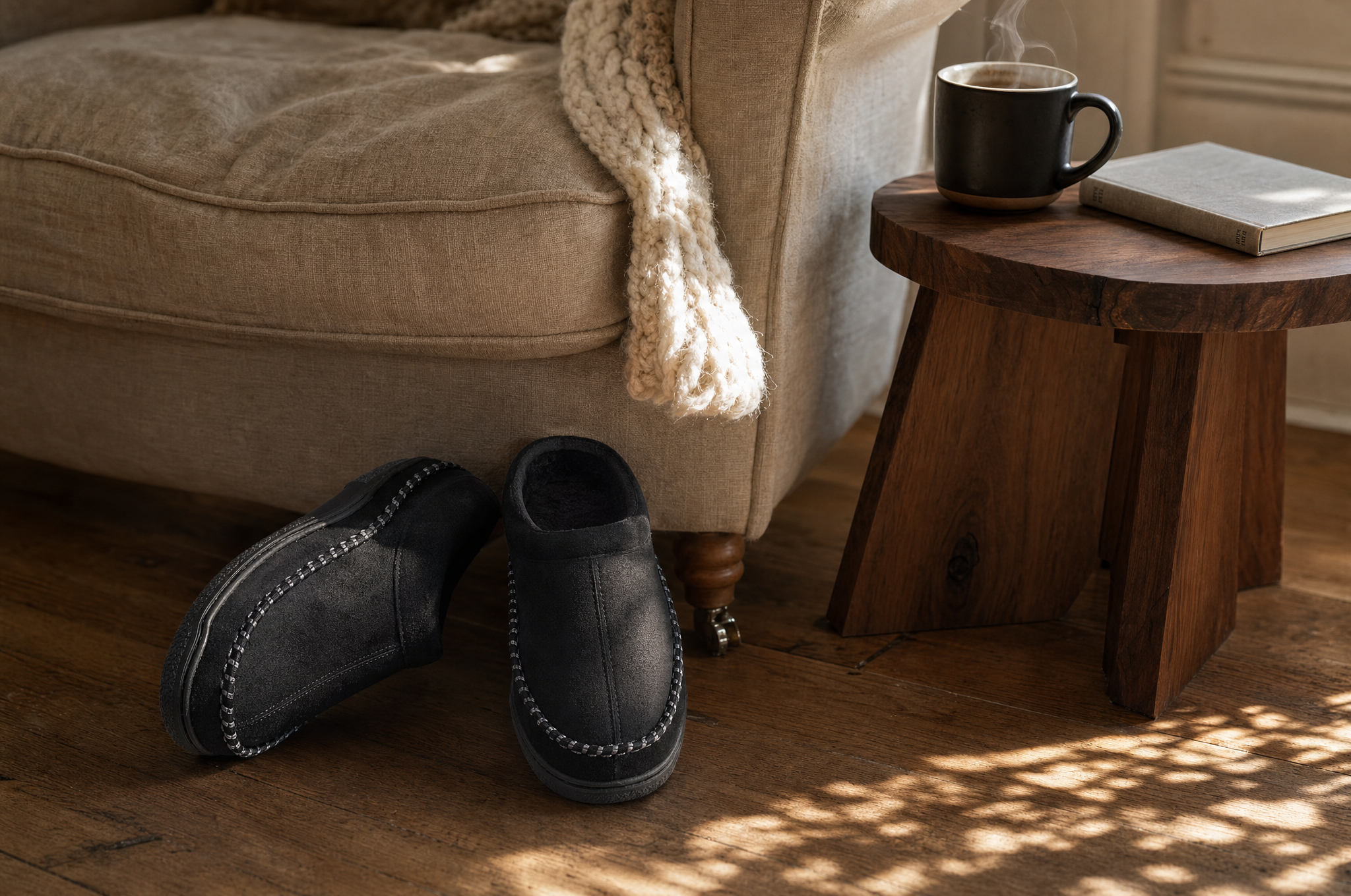 Black fleece-lined moccasin slippers beside an armchair with a steaming mug on a side table
