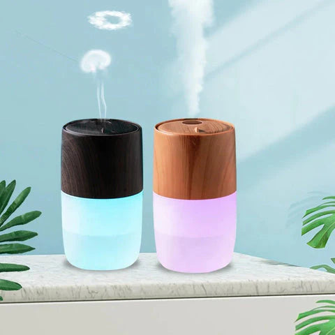 Black and woodgrain jellyfish-inspired humidifiers on a table, emitting mist with ambient LED lighting, surrounded by green leaves.