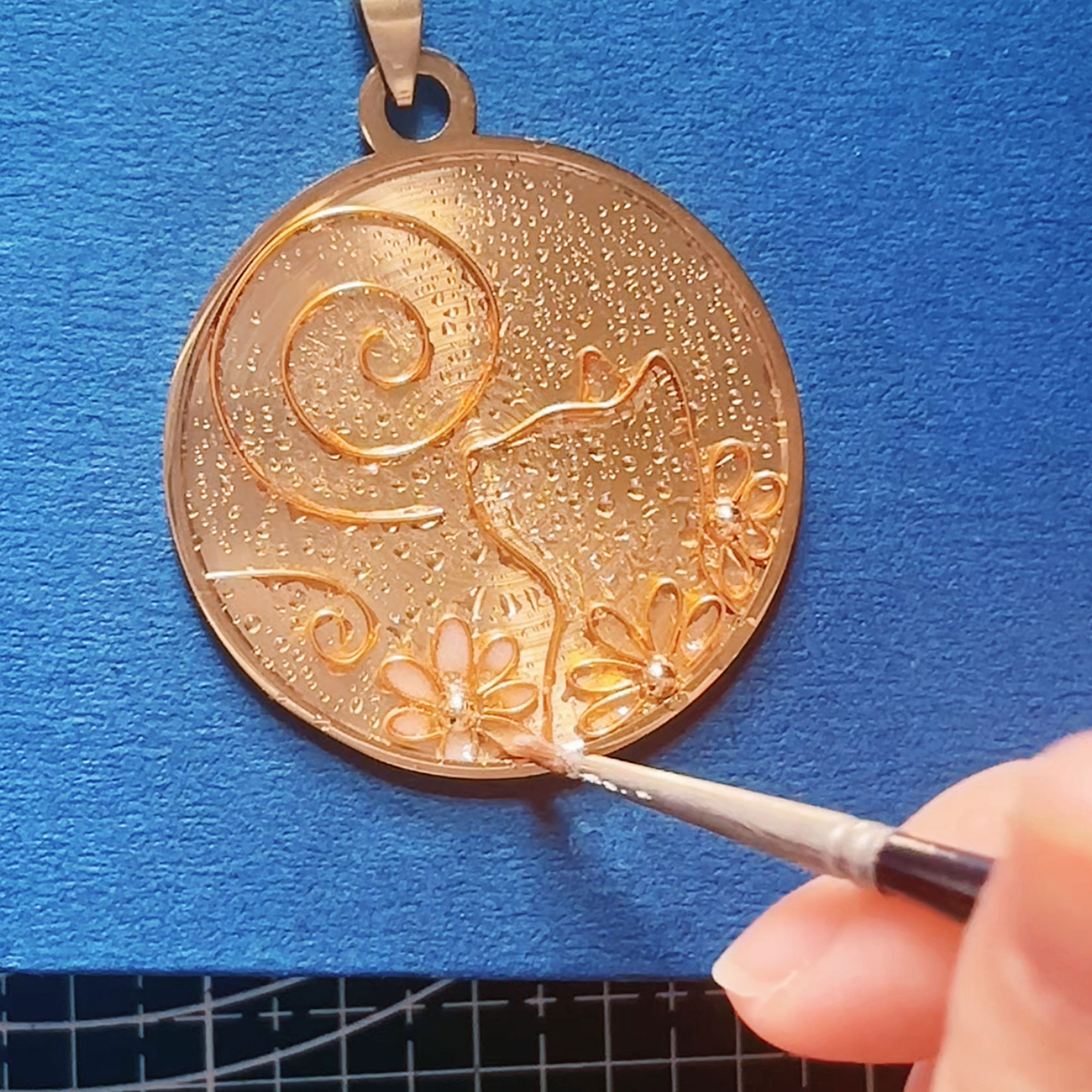 Close-up of artisan hands using a fine needle tool to meticulously hand-paint and hand-pour colorful enamel sand into the tiny wire compartments of a pendant.