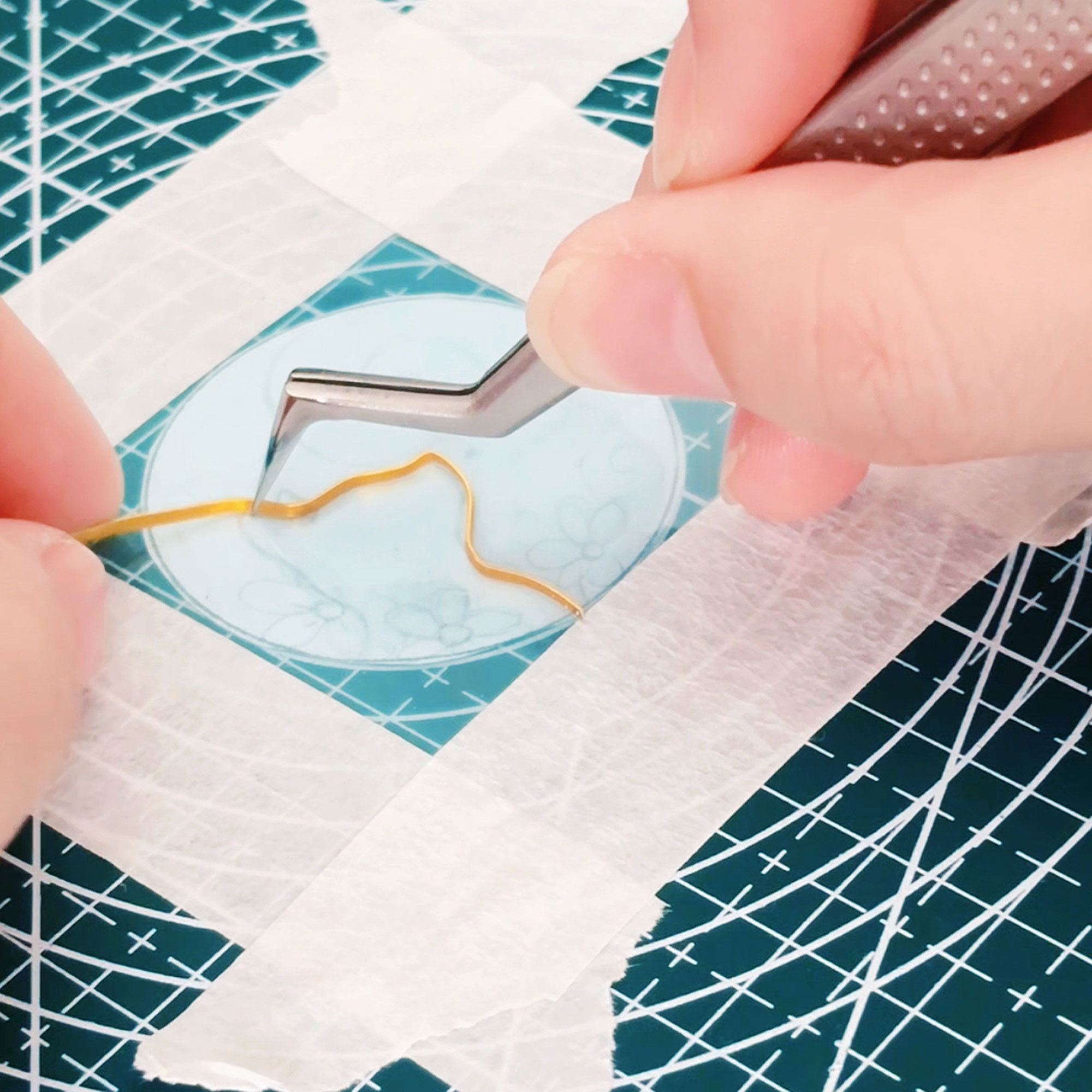 Artisan hands using tweezers to carefully bend and shape a thin gold wire into the outline of a cat on a sticky film. This traditional Cloisonné wirework technique is the foundation of my custom handmade jewelry.