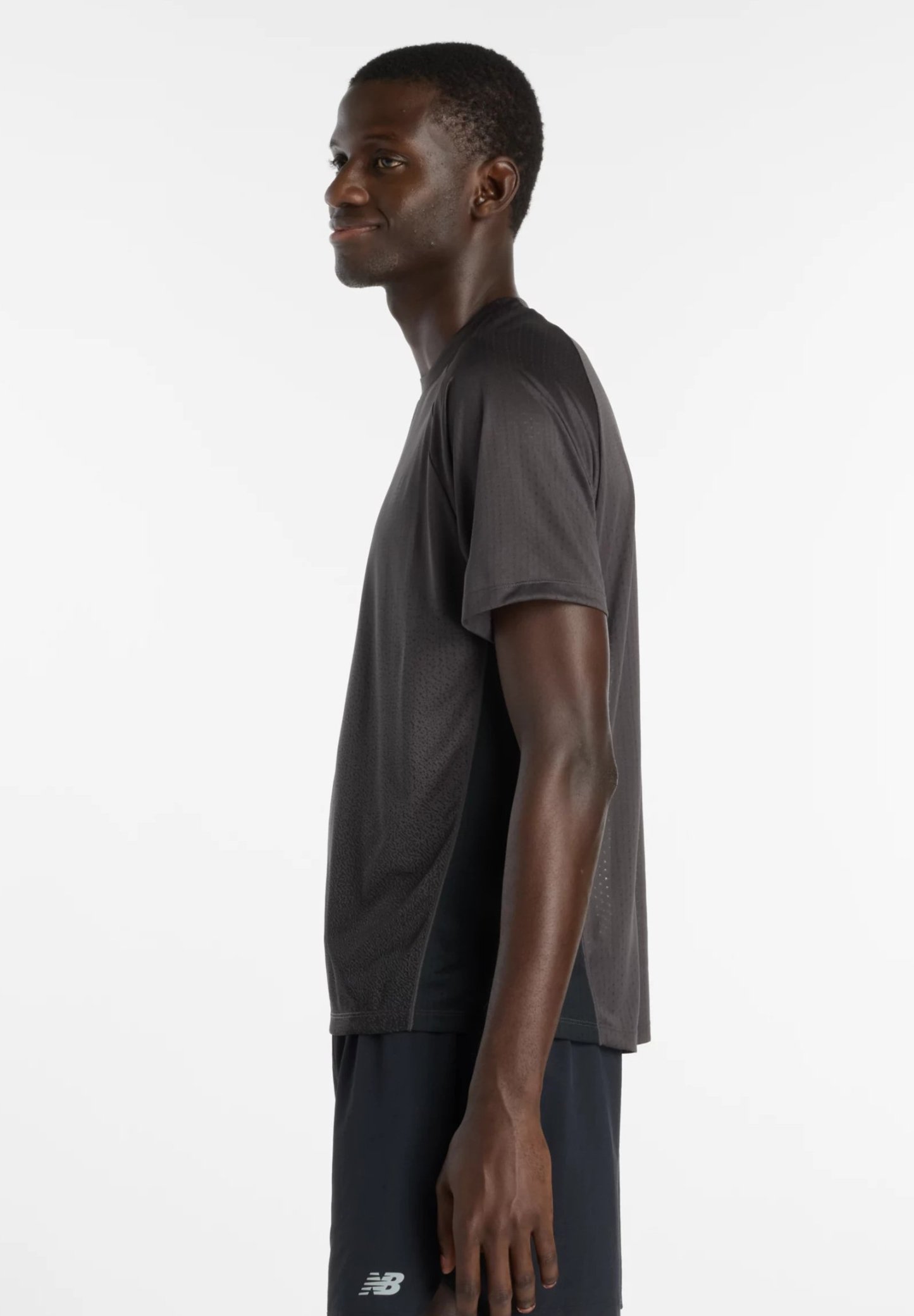 Joven con camiseta deportiva negra y pantalones cortos, de perfil sobre un fondo blanco liso, sonriendo ligeramente.