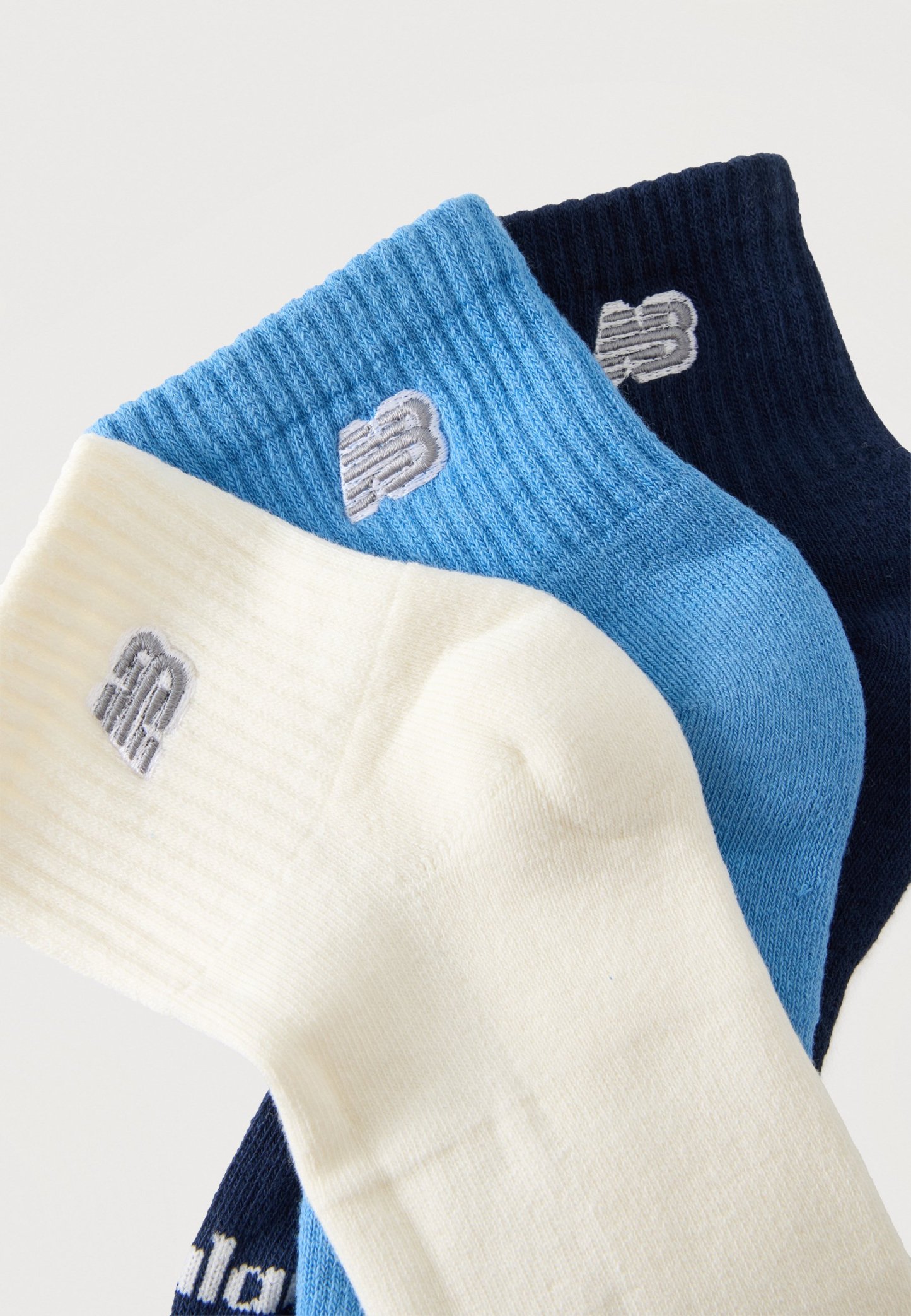 Tres pares de calcetines tobilleros acanalados en blanco, azul claro y azul marino, cada uno con un pequeño logo bordado cerca del puño.