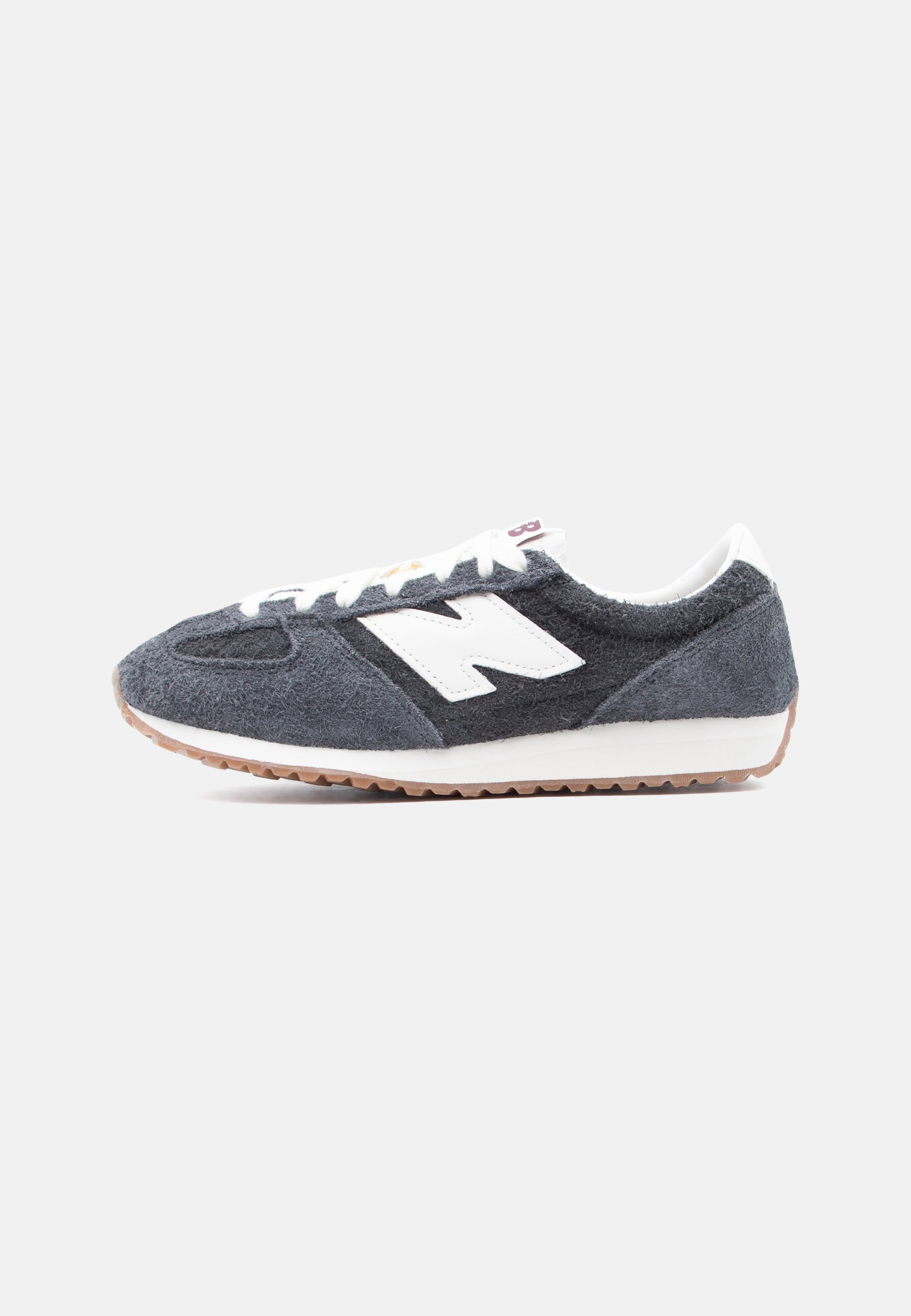 Zapatilla de ante gris oscuro y negro con cordones blancos, logo "N" blanco en el lateral y suela de goma marrón claro.