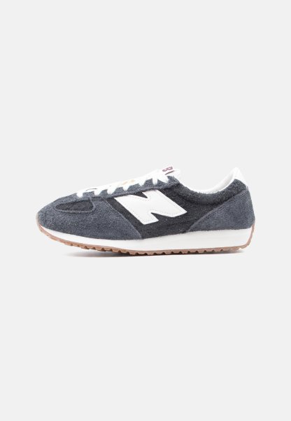 Zapatilla de ante gris oscuro y negro con cordones blancos, logo "N" blanco en el lateral y suela de goma marrón claro.