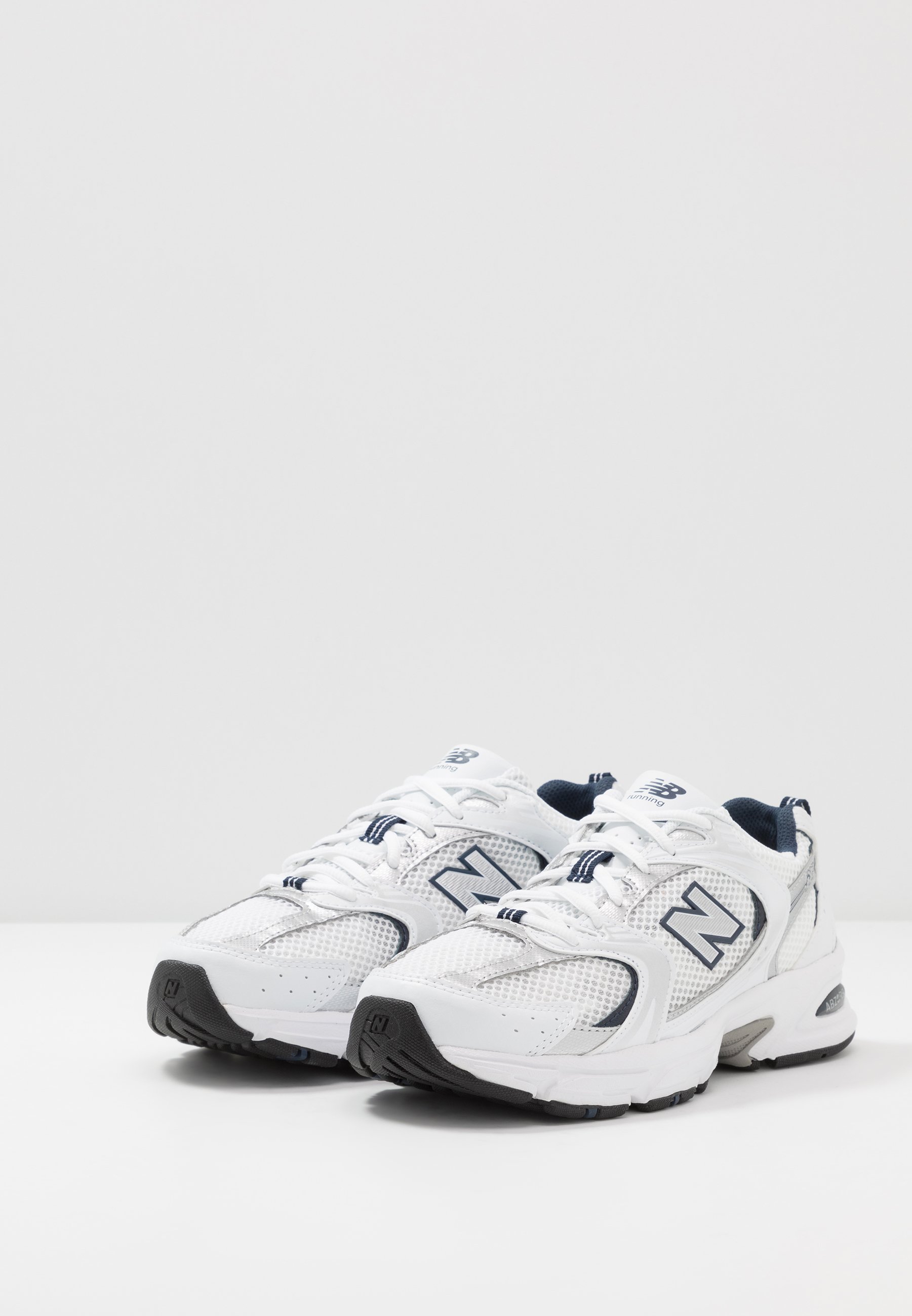 Zapatillas deportivas blancas con material de malla y sintético, que presentan una suela exterior gris, acentos en azul oscuro y un prominente logo "N" en el lateral.