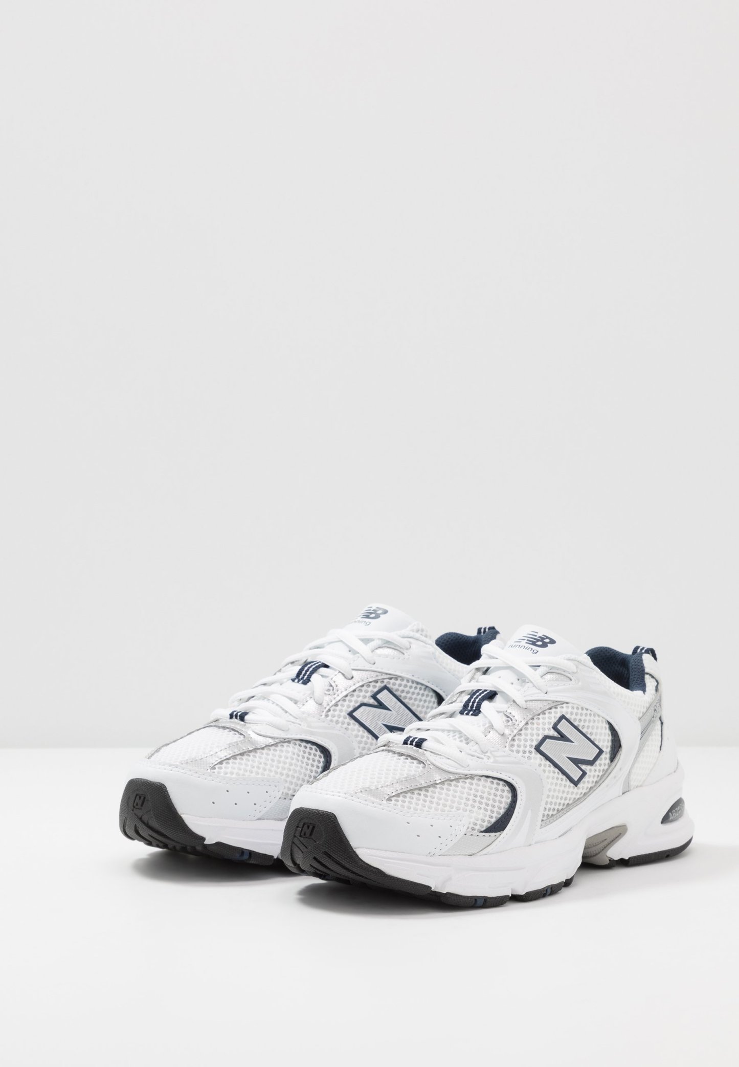 Zapatillas deportivas blancas con material de malla y sintético, que presentan una suela exterior gris, acentos en azul oscuro y un prominente logo "N" en el lateral.