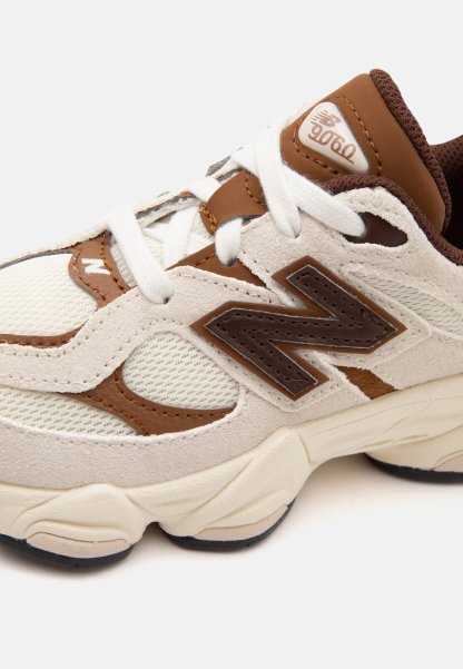 Primer plano de una zapatilla New Balance 9060 beige y marrón con cordones blancos y suela gruesa sobre fondo claro.