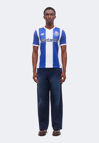 Hombre de pie mirando hacia adelante, con una camiseta de fútbol a rayas azules y blancas y vaqueros anchos azul oscuro, sobre un fondo claro y sencillo.