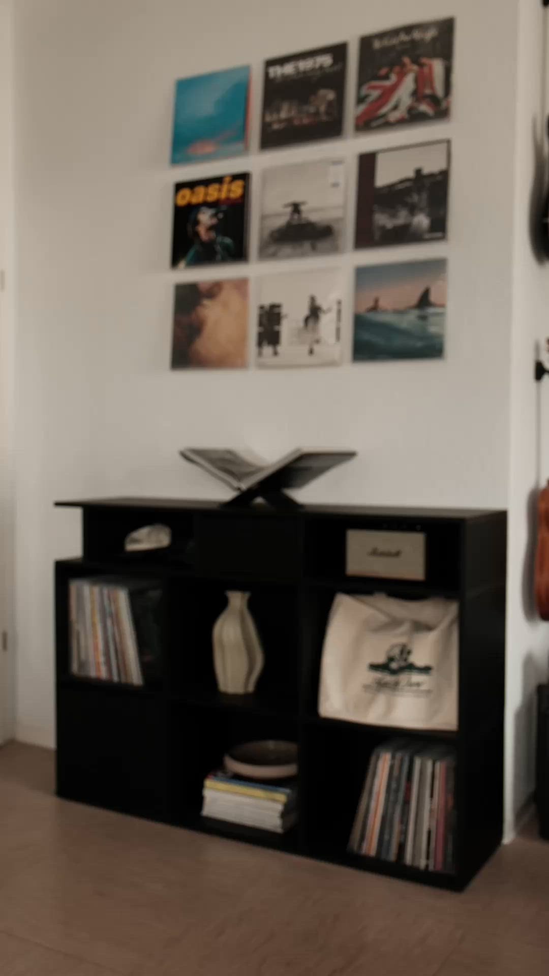 Unidad de almacenamiento de madera negra con múltiples cubículos abiertos, que exhibe discos de vinilo, un jarrón y una bolsa. Los discos montados en la pared arriba muestran varias portadas de álbumes.