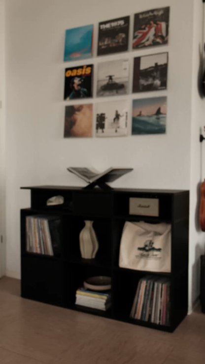 Unidad de almacenamiento de madera negra con múltiples cubículos abiertos, que exhibe discos de vinilo, un jarrón y una bolsa. Los discos montados en la pared arriba muestran varias portadas de álbumes.