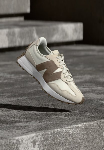 Zapatilla beige y blanca con detalles en ante marrón, cordones blancos y suela texturizada, situada sobre una superficie de hormigón y escaleras al fondo.