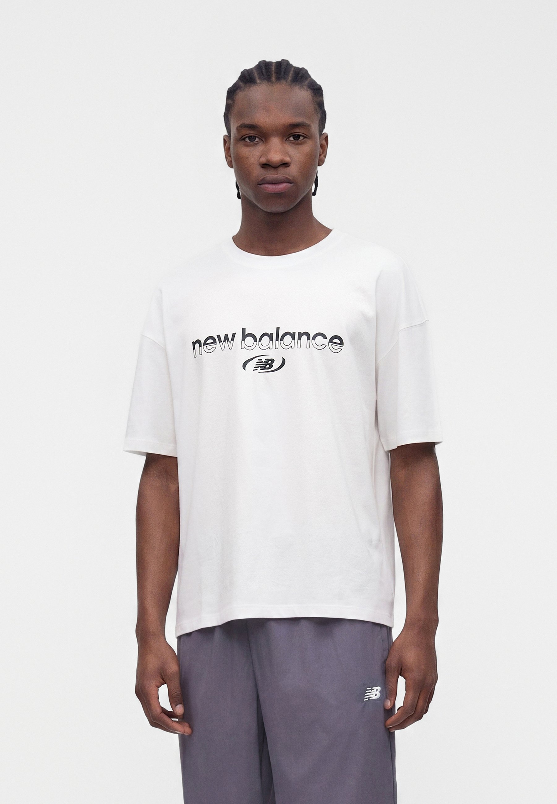 Joven con el pelo trenzado, vestido con una camiseta blanca de New Balance y pantalones grises, de pie frente a un fondo blanco liso.