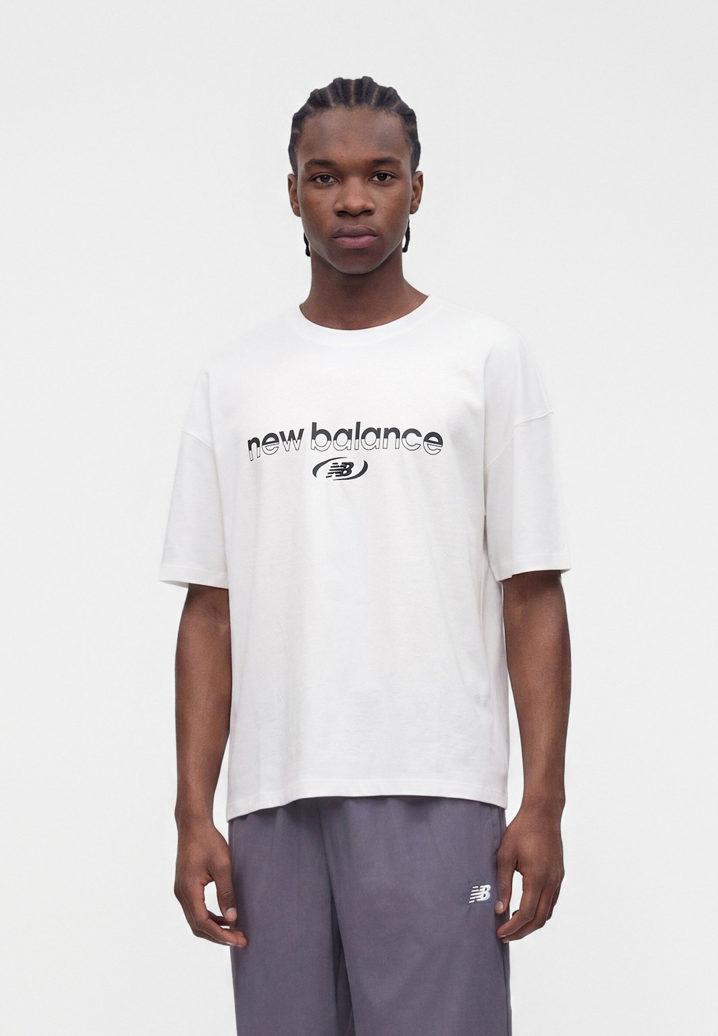 Joven con el pelo trenzado, vestido con una camiseta blanca de New Balance y pantalones grises, de pie frente a un fondo blanco liso.