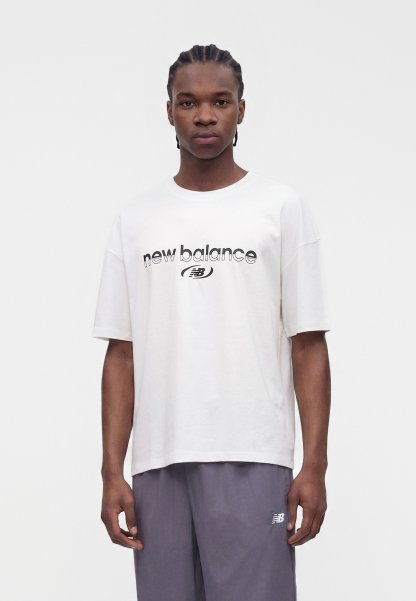 Joven con el pelo trenzado, vestido con una camiseta blanca de New Balance y pantalones grises, de pie frente a un fondo blanco liso.