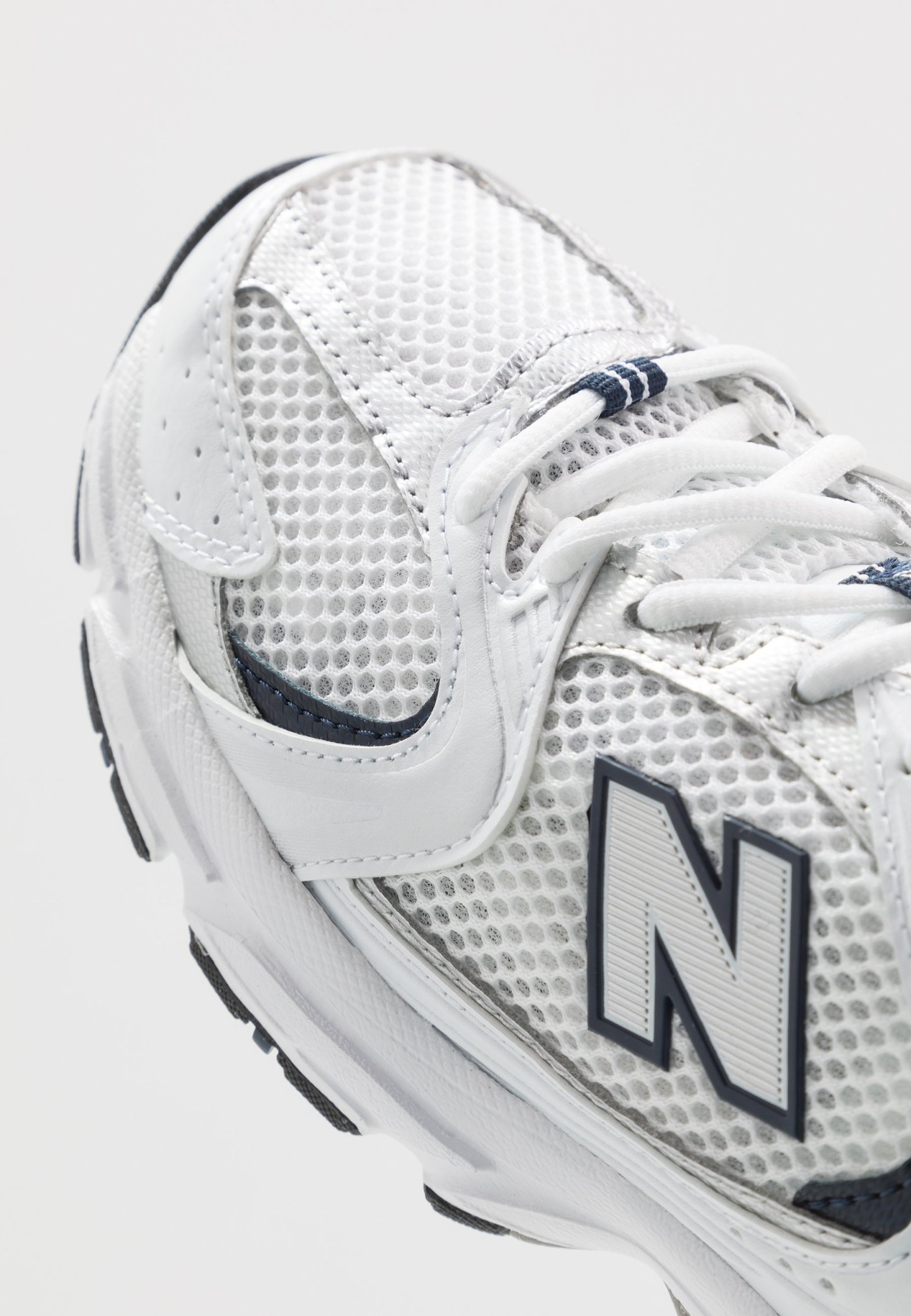 Zapatilla deportiva blanca con parte superior de malla, acentos de cuero y detalles en azul marino. Forma redondeada con un prominente logo 'N' y superficie texturizada.