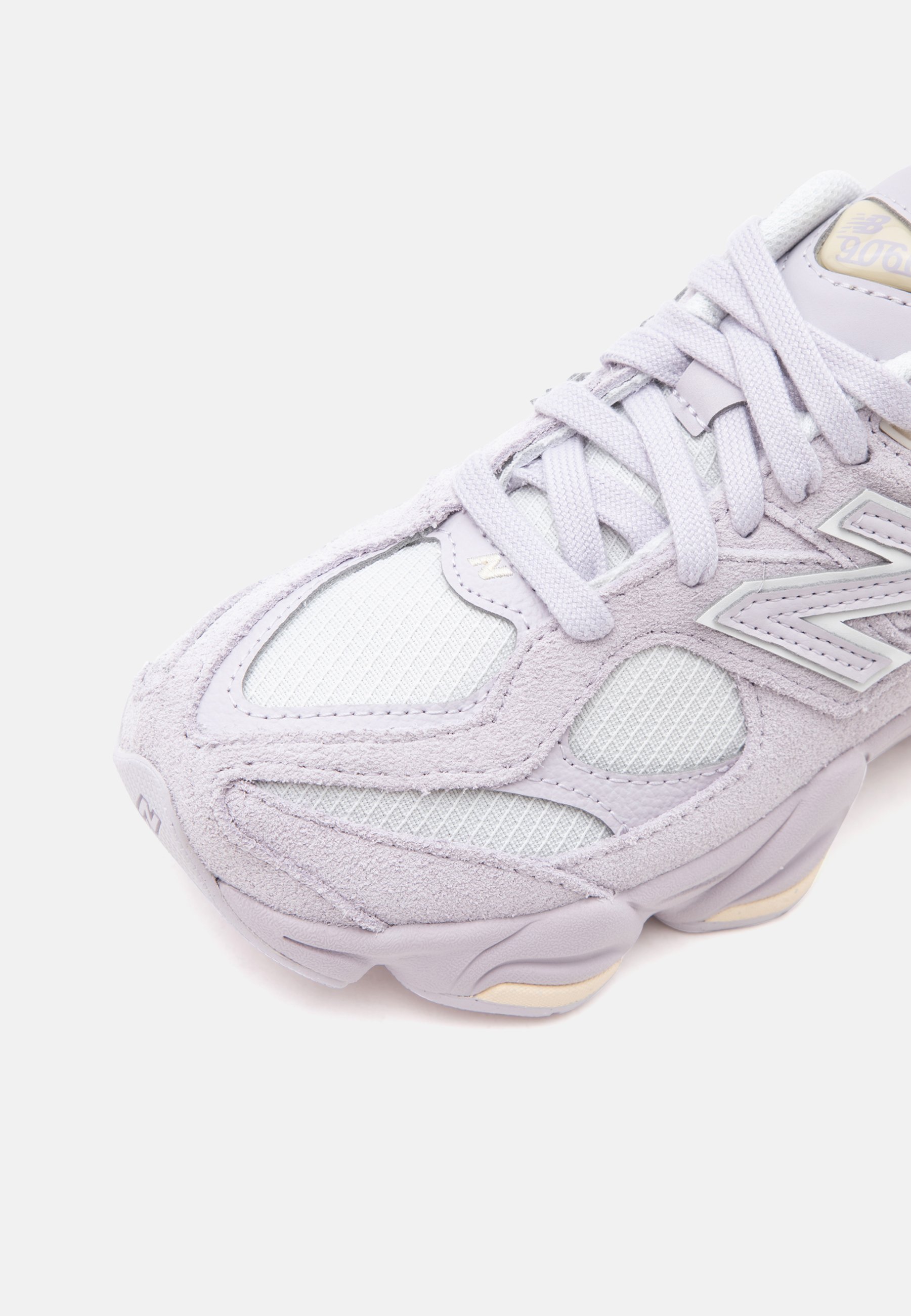 Primer plano de una zapatilla deportiva gris claro con paneles de malla, superposiciones de ante y cordones, con un logo de la marca en el lateral.