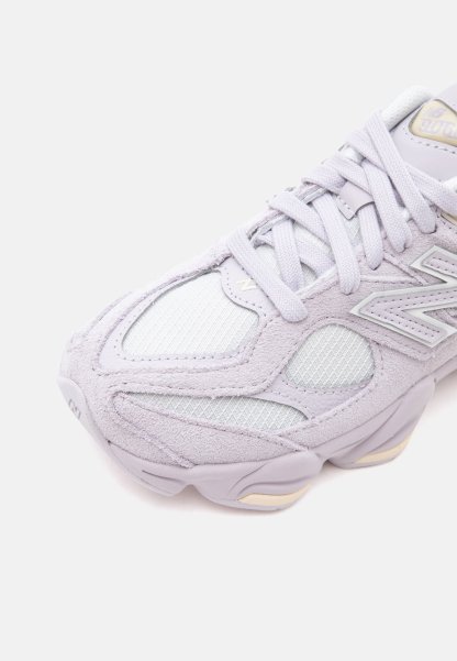 Primer plano de una zapatilla deportiva gris claro con paneles de malla, superposiciones de ante y cordones, con un logo de la marca en el lateral.
