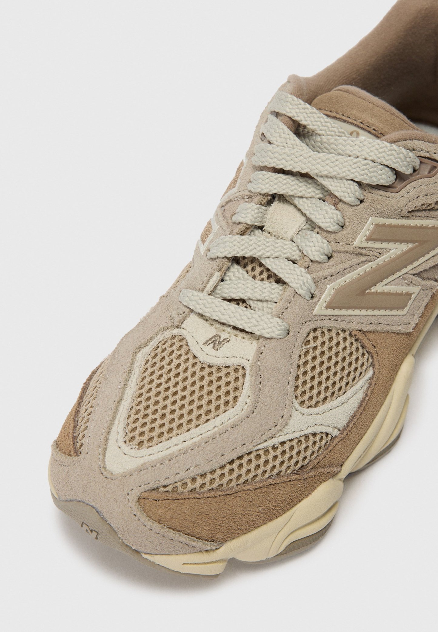 Zapatilla atlética beige con paneles de malla y acentos de ante. Presenta cordones de colores claros y suela acolchada. Incluye un gran detalle del logo en el lateral.