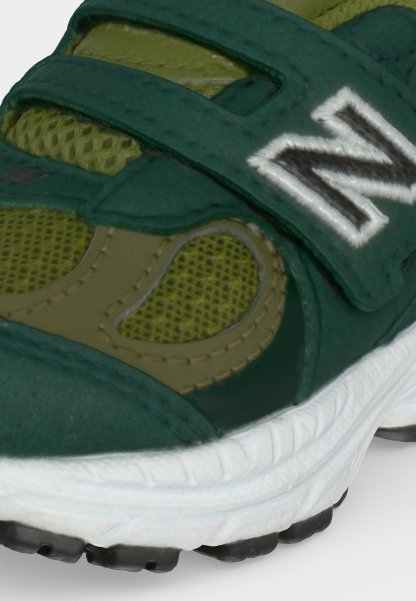 Primer plano de una zapatilla verde y oliva con tejido de malla, una correa de velcro con un "N," blanco bordado y una suela blanca texturizada.