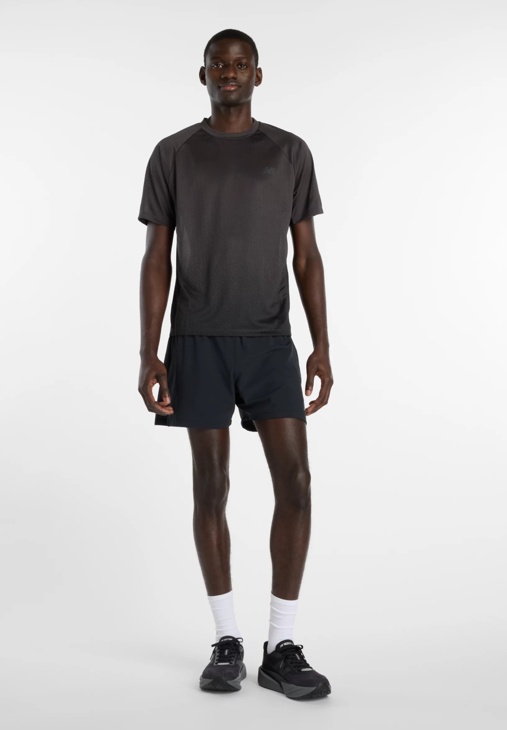 Hombre atlético de pie, vistiendo camiseta deportiva negra, pantalones cortos negros, calcetines blancos hasta la pantorrilla y zapatos de correr negros sobre un fondo blanco liso.