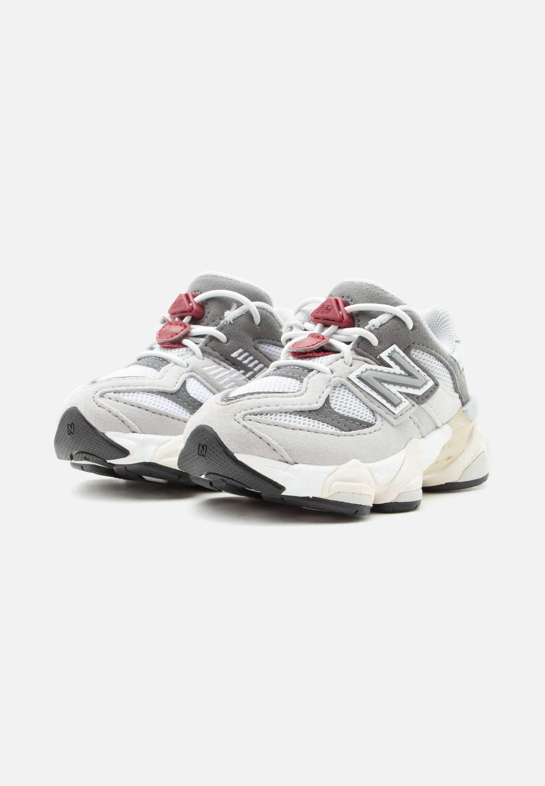 Par de zapatillas New Balance en gris y blanco con paneles de malla y ante, sujetadores de cordones rojos y suelas de goma negras inclinadas hacia adelante.