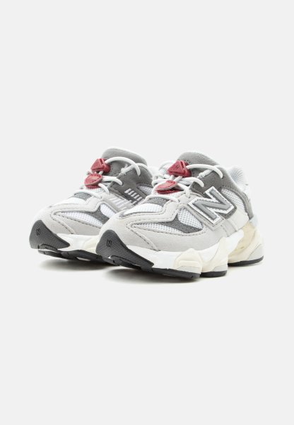 Par de zapatillas New Balance en gris y blanco con paneles de malla y ante, sujetadores de cordones rojos y suelas de goma negras inclinadas hacia adelante.