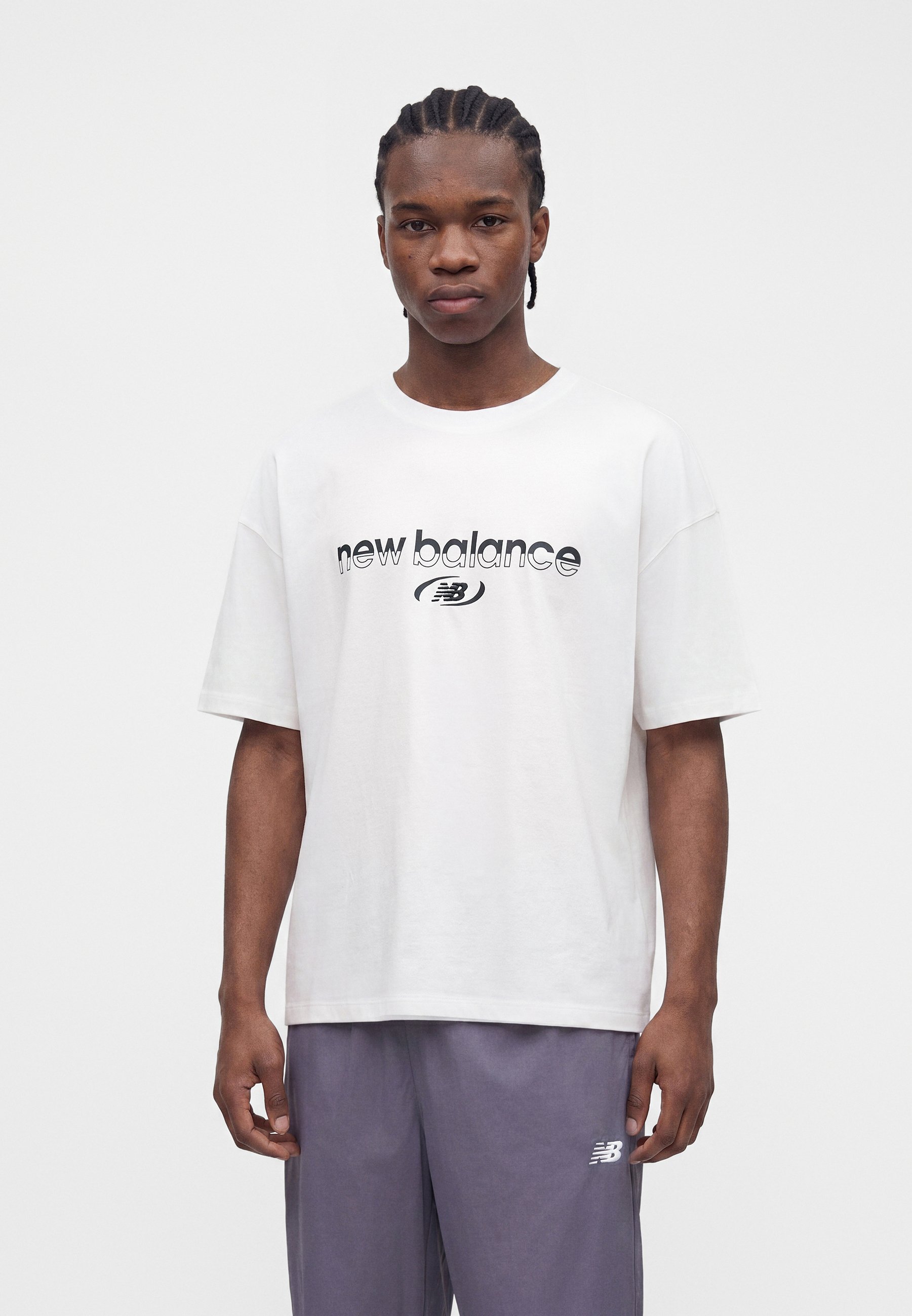 Joven con una camiseta blanca de "new balance" y pantalones grises con el logo "NB", de pie frente a un fondo blanco liso.