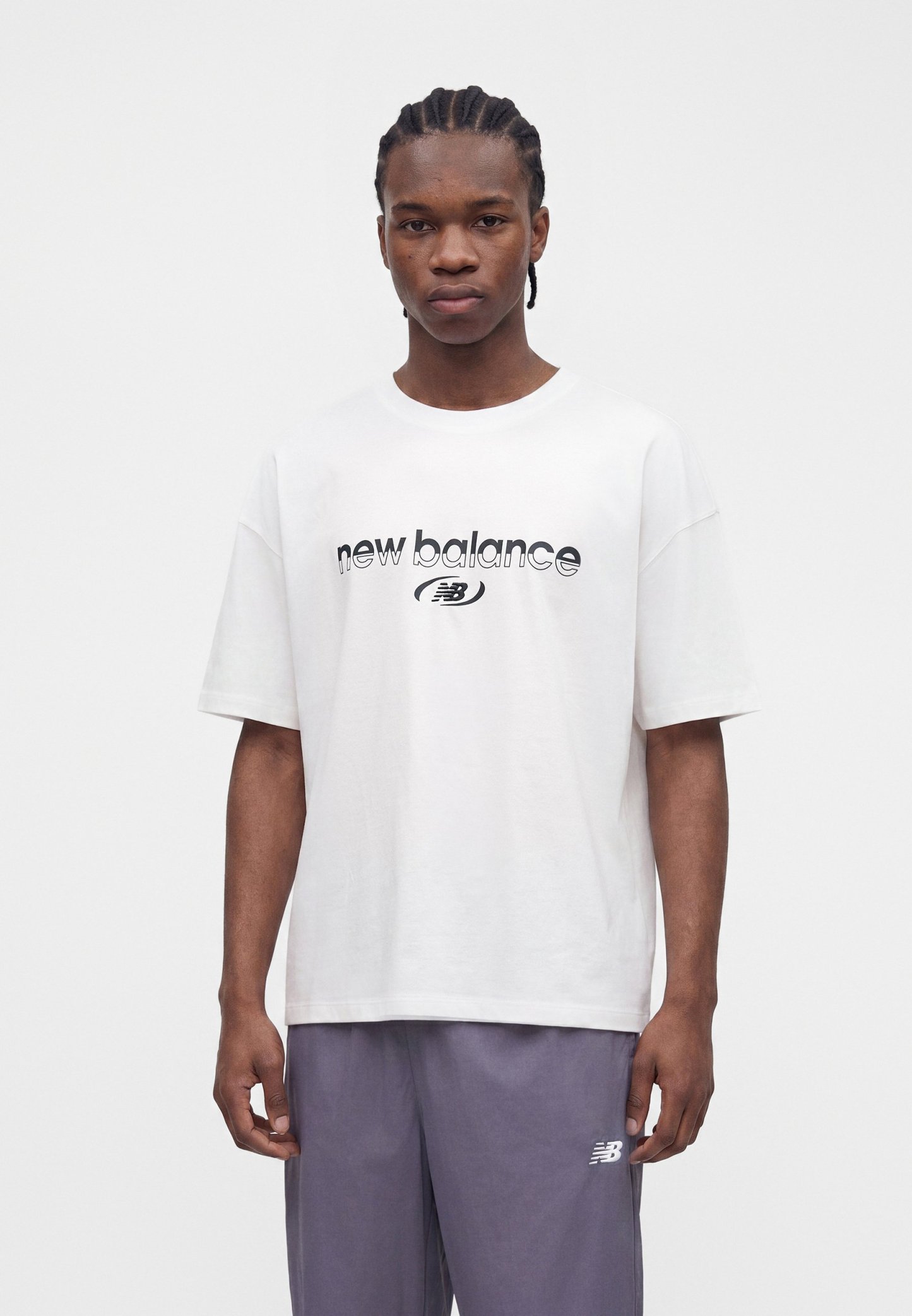 Joven con una camiseta blanca de "new balance" y pantalones grises con el logo "NB", de pie frente a un fondo blanco liso.