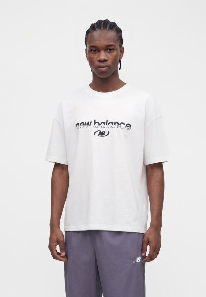 Joven con una camiseta blanca de "new balance" y pantalones grises con el logo "NB", de pie frente a un fondo blanco liso.