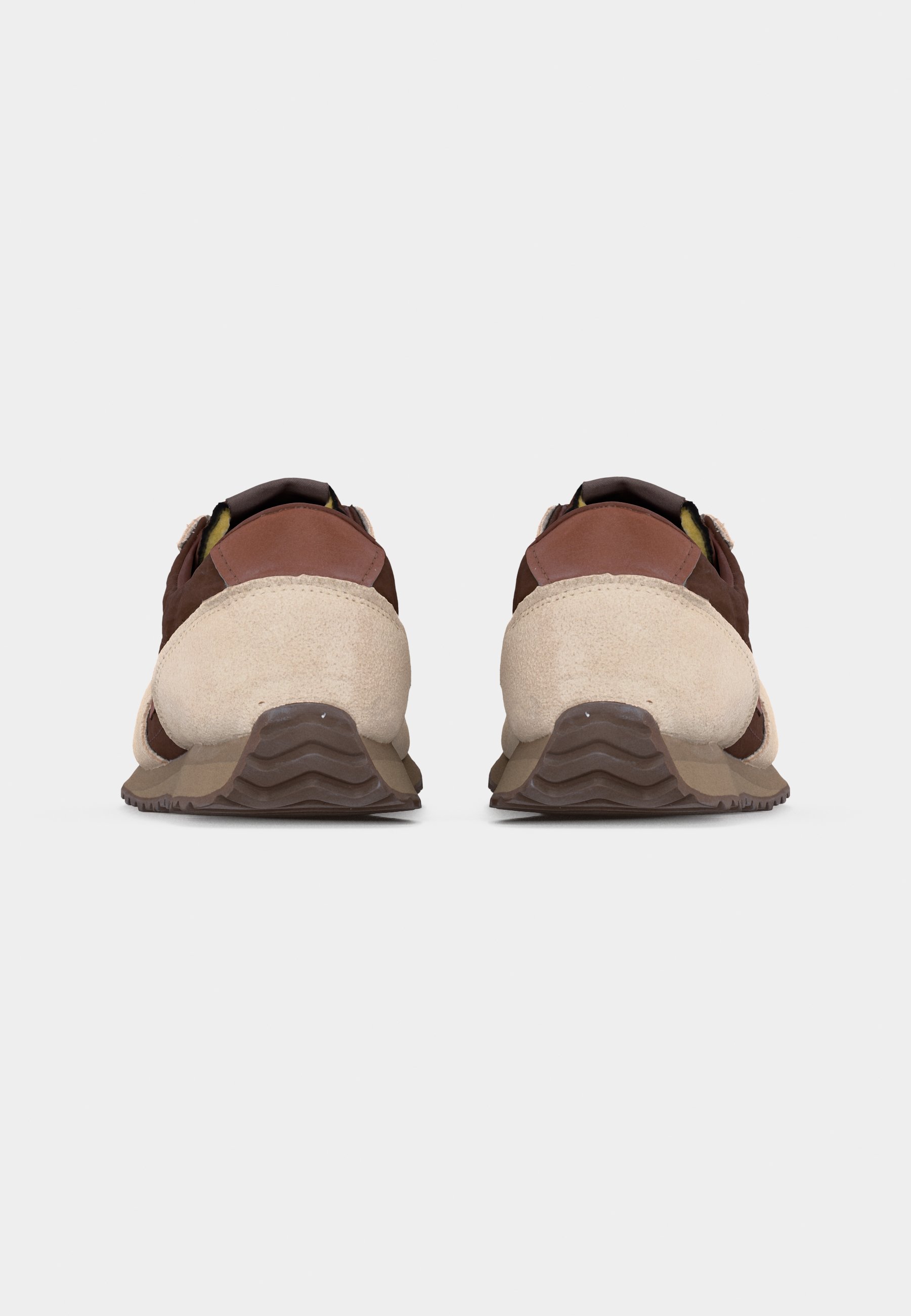 Par de zapatillas marrones y beige vistas desde atrás sobre un fondo liso gris claro, destacando las suelas texturizadas y los detalles en ante.