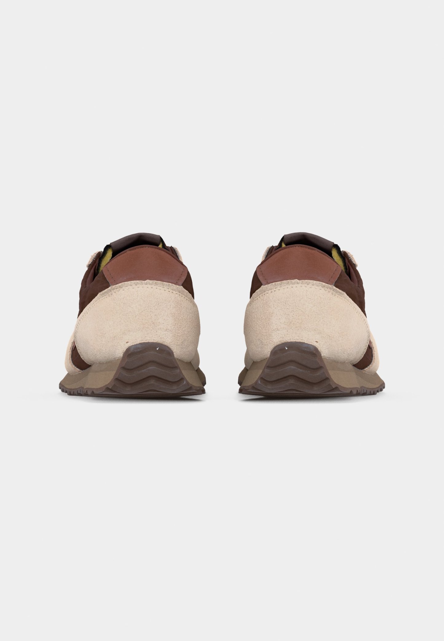 Par de zapatillas marrones y beige vistas desde atrás sobre un fondo liso gris claro, destacando las suelas texturizadas y los detalles en ante.