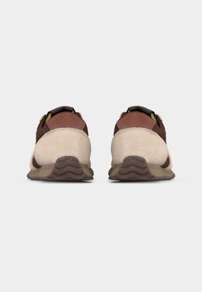 Par de zapatillas marrones y beige vistas desde atrás sobre un fondo liso gris claro, destacando las suelas texturizadas y los detalles en ante.