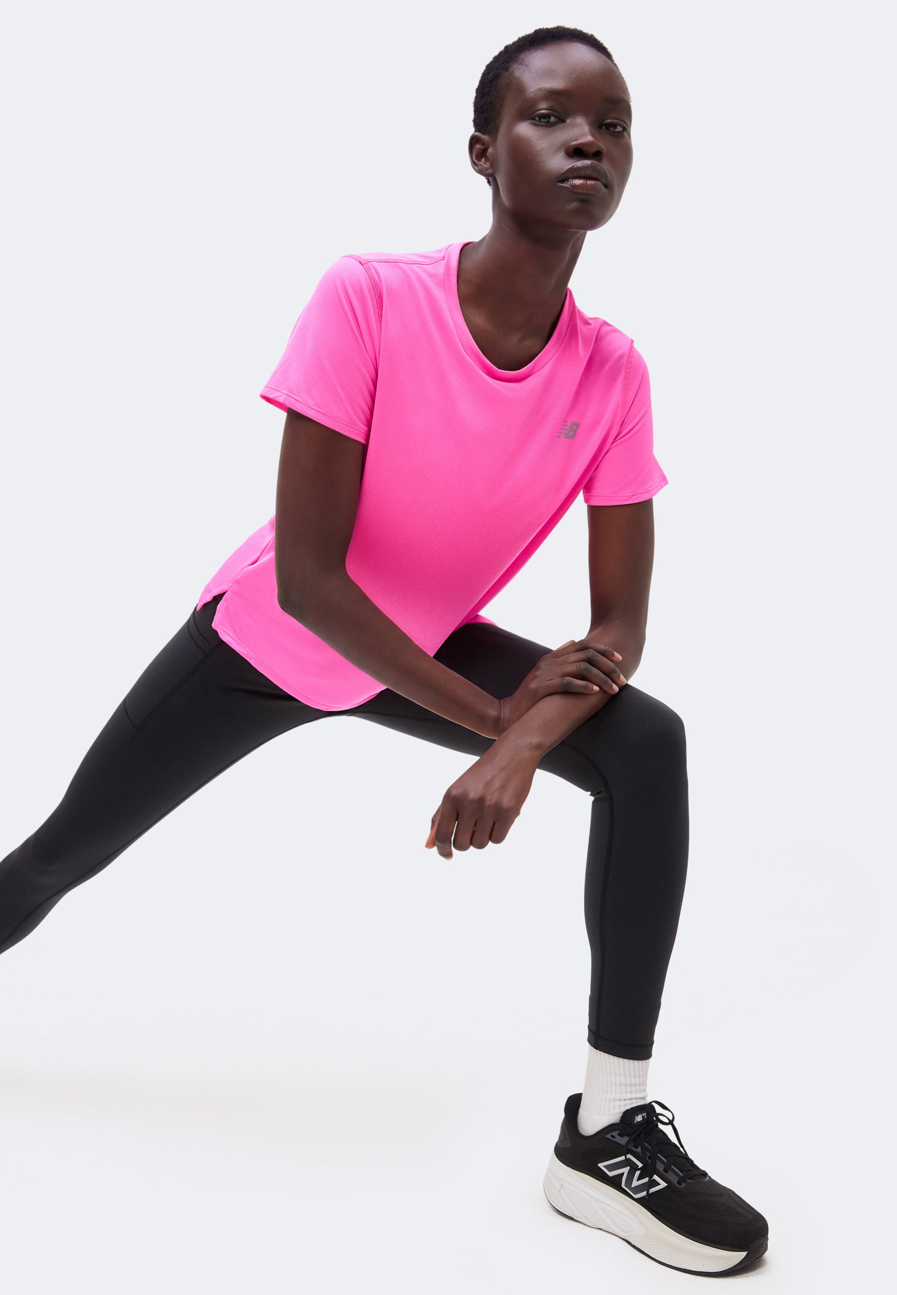 Mujer con camiseta rosa brillante, leggings negros y zapatillas negras estira una pierna hacia adelante con las manos apoyadas en la rodilla doblada.