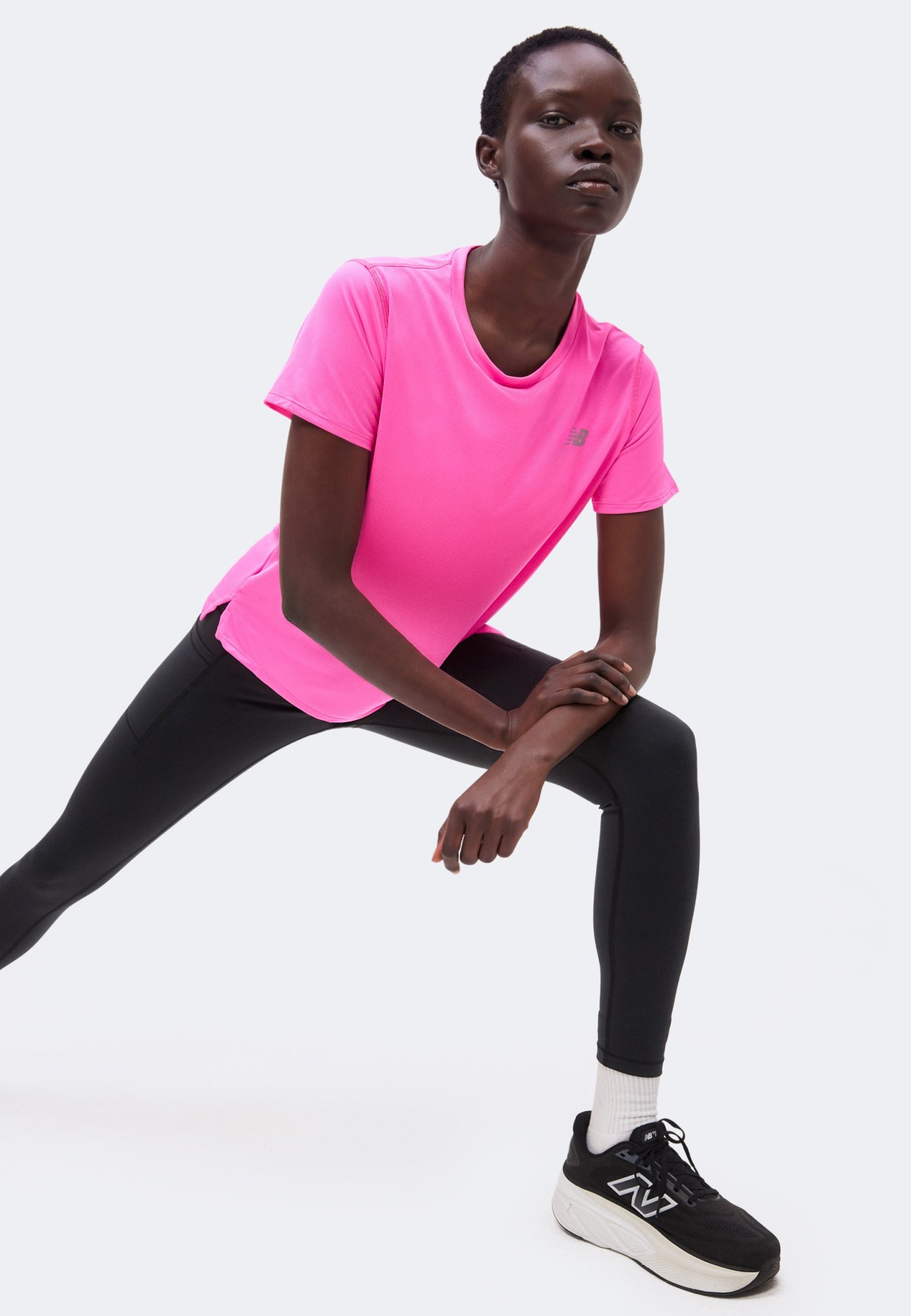 Mujer con camiseta rosa brillante, leggings negros y zapatillas negras estira una pierna hacia adelante con las manos apoyadas en la rodilla doblada.