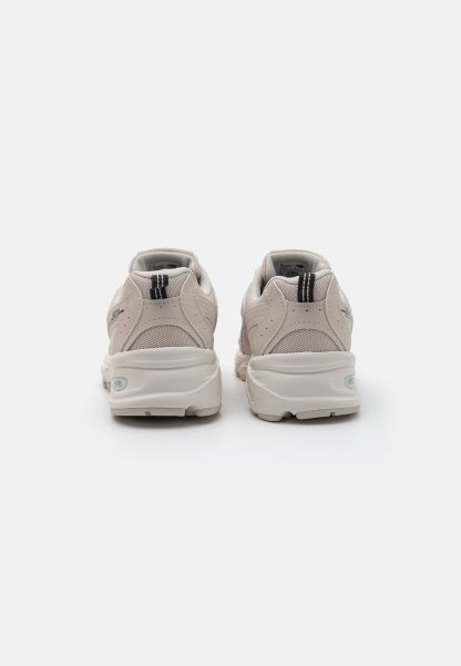 Zapatillas de deporte color beige claro, hechas de material de malla y goma, con forma redondeada, superposiciones texturizadas y un acento de franja negra en el talón.