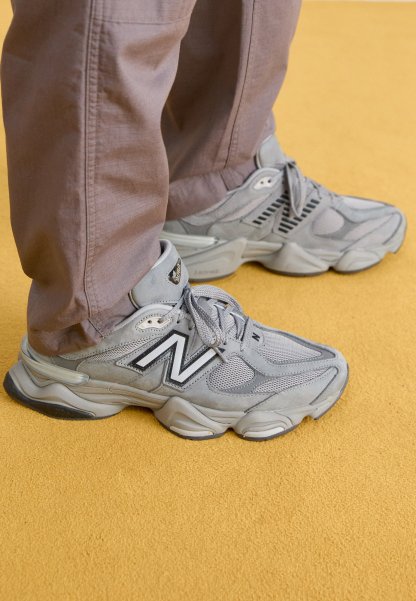 Persona con zapatillas New Balance grises con suelas gruesas y pantalones marrones, de pie sobre un suelo alfombrado amarillo.