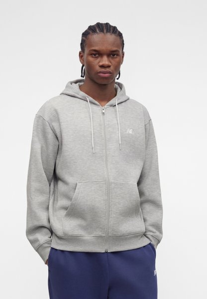 Joven con trenzas, vestido con una sudadera con cremallera gris claro y pantalones azul oscuro sobre un fondo blanco liso.