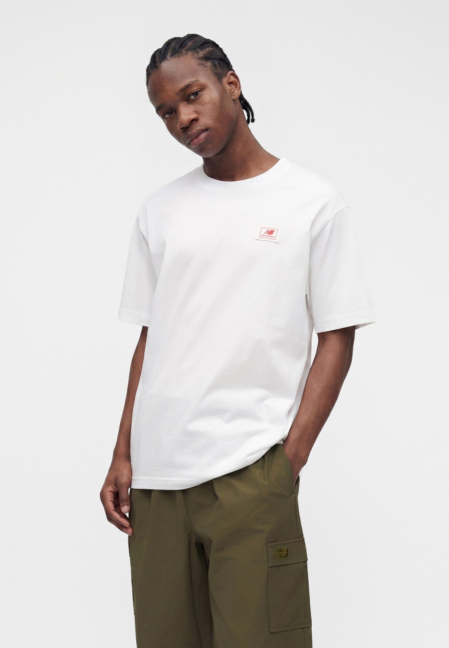 Joven con cabello trenzado que lleva una camiseta blanca de New Balance y pantalones cargo verde oliva, de pie con una mano en el bolsillo.